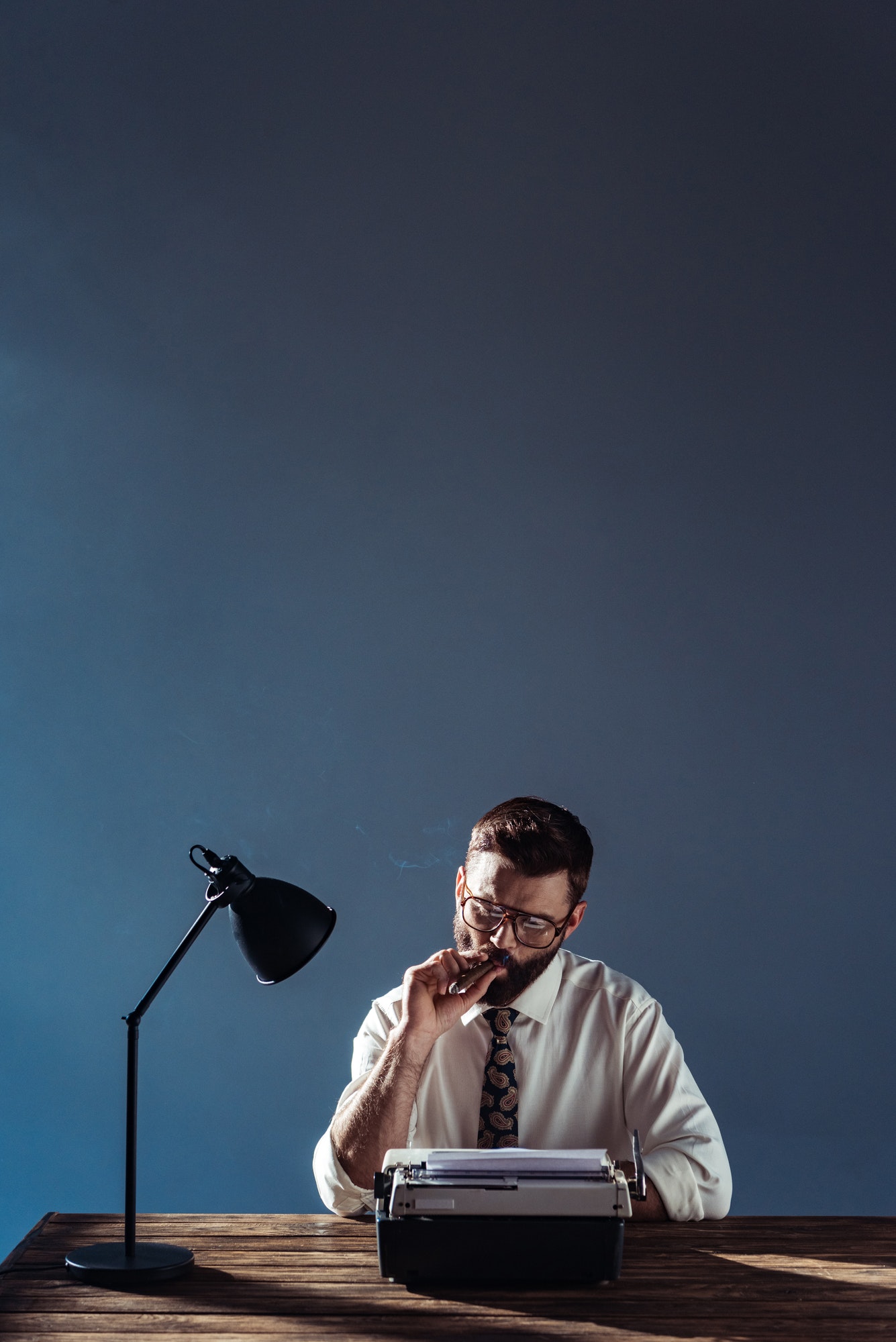 handsome journalist sitting at table with lamp and retro typewriter and smoking on grey background